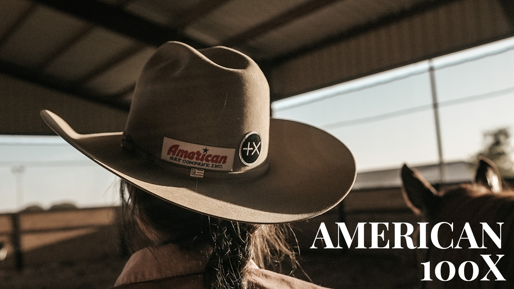 A man wearing an American Hat Company 100x felt hat is seen from behind looking off at a dirt ground with overcast skies.