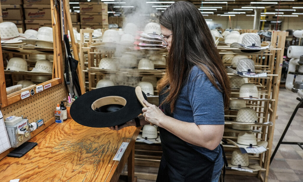 photo of a female employee with long, dark hair working and shaping a black felt hat with steam, a mirror. and racks of straw hats in the background
