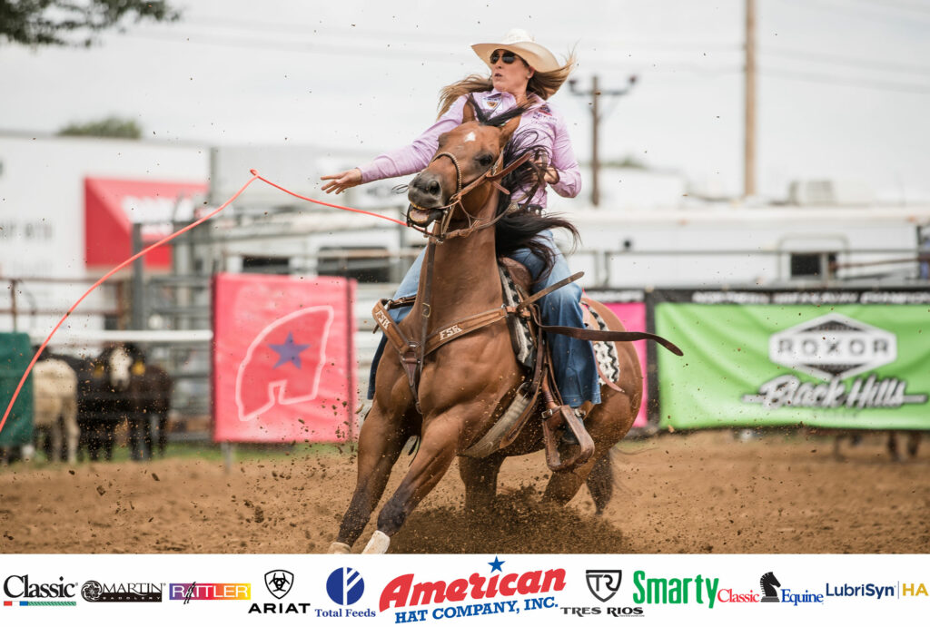 Jackie Crawford rides a brown horse with a rope in her hand. She wears a pink button-up shirt and denim jeans. She has a straw cowboy hat on her head.