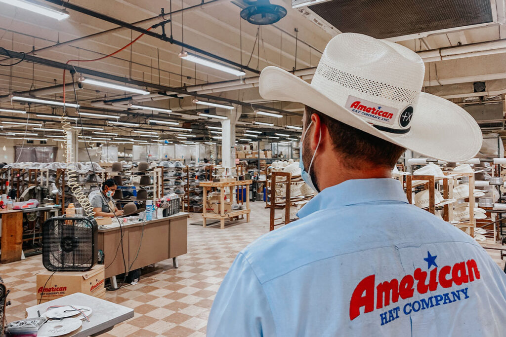 A man is shown from behind wearing a denim button-up shirt with American Hat Company embroidered on the back. He is walking into the work area where they make American Hat Company hats.