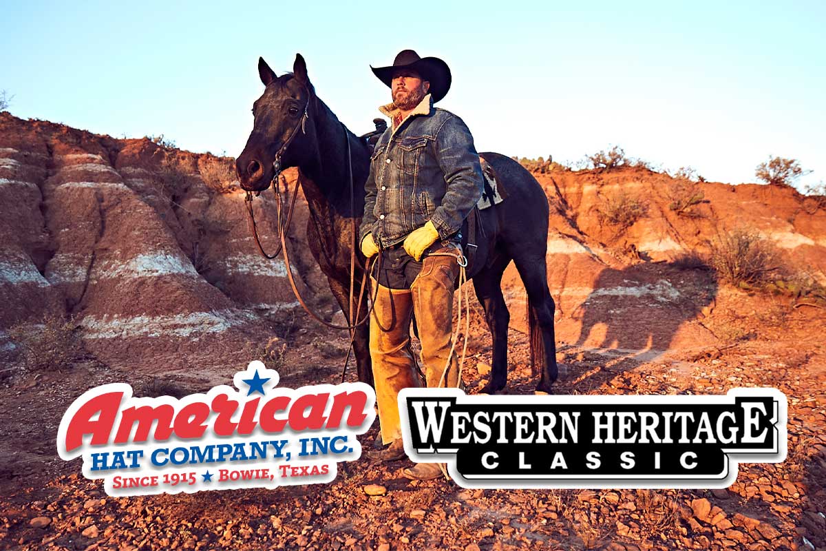 A cowboy and a horse are standing with rugged terrain in the background. The cowboy wears chaps, a denim jacket, and a black felt hat. The American Hat Company logo and the Western Heritage Classic logo are overlaid along the bottom of the photo.