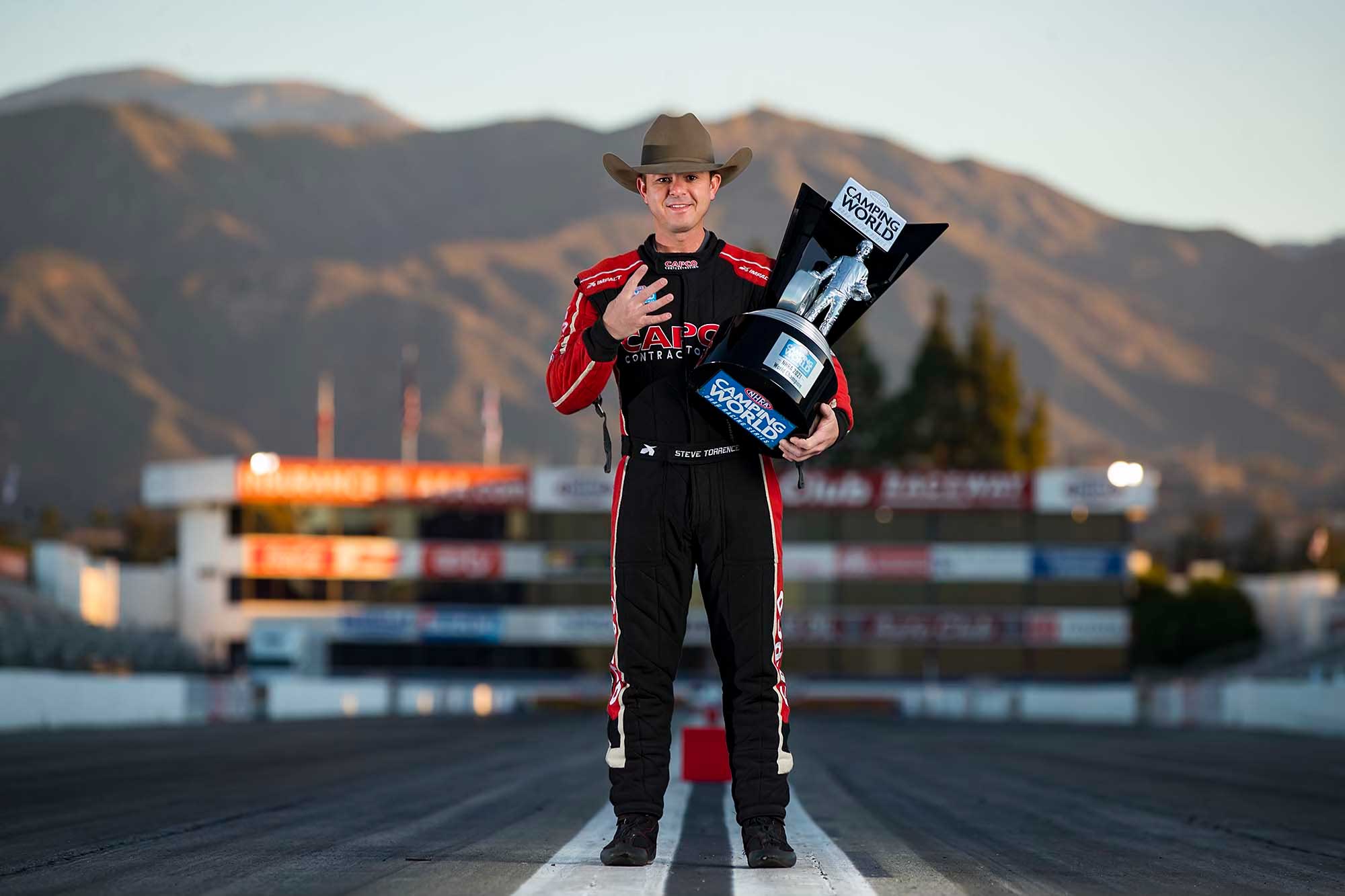 Steve Torrence wears a black and red jumpsuit while holding a large trophy.