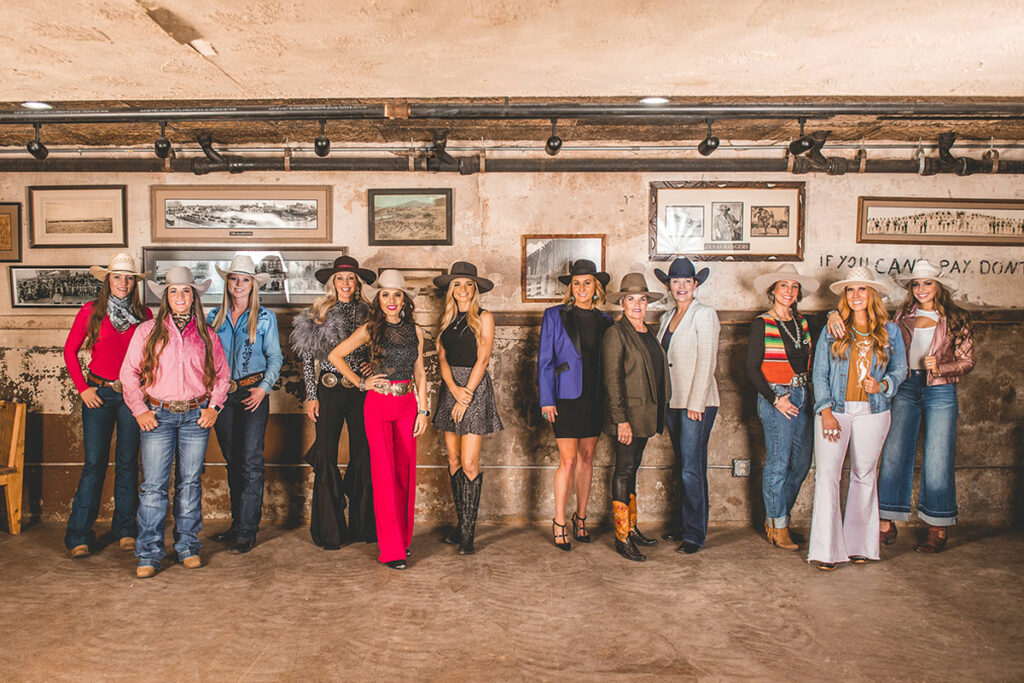 A large group of women stand in a room with rustic tan walls and floor. They are all wearing fashion hats by American Hat Company.